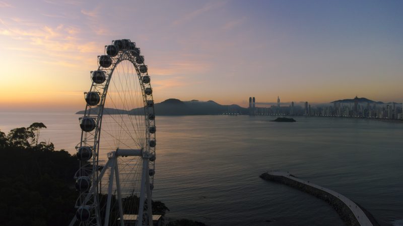 Foto da Roda Gigante de Balneário Camboriú. Ela está no canto esquerdo da foto, ao lado da vegetação do Morro da Rainha. No canto direito, o molhe do Pontal Norte. Ao centro e pelo horizonte, o mar, prédios na orla da Praia Central e um morro verde. O céu está em tons de azul e laranja. 