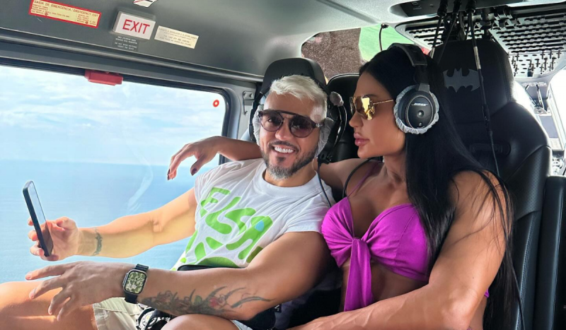 Belo e Gracyanne Barbosa voando em direção ao Cruzeiro de Neymar - Homem de cabelos loiros e mulher de cabelos pretos sentados dentro de helicóptero.
