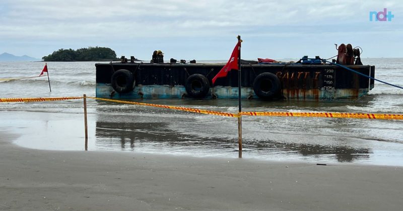 Imagem mostra barcaça encalhada na praia Central, bem de perto, isolada por fica de segurança