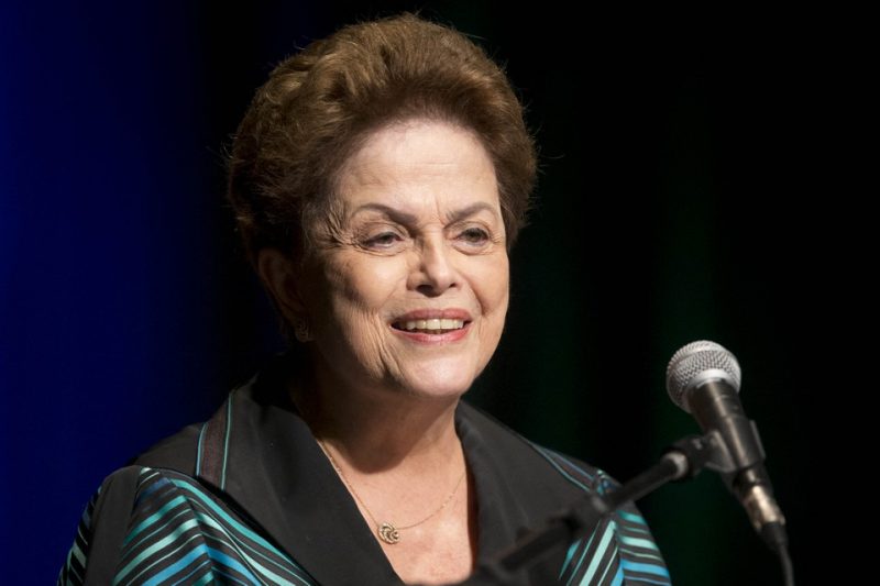 Dilma Rousseff, mulher branca com cabelos curtos sorrindo sentada em frente a um microfone 