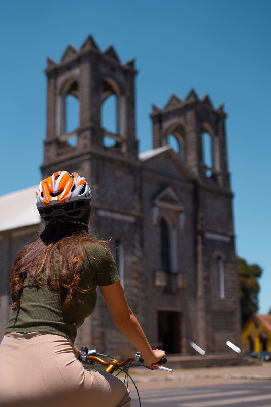Na imagem, uma moça de costas equipada para andar de bicicleta e São Joaquim, na frente de um ponto turístico da cidade