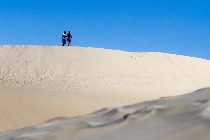 Foto de uma duna de areia clara da Praia dos Ingleses. Em cima da duna, há um homem e uma mulher. O céu está bem azul. 