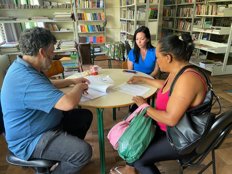 Foto mostra mesa eleitoral com dois mesários e uma mãe pegando as cédulas de votação na escola EEB Jurema Cavallazzi 