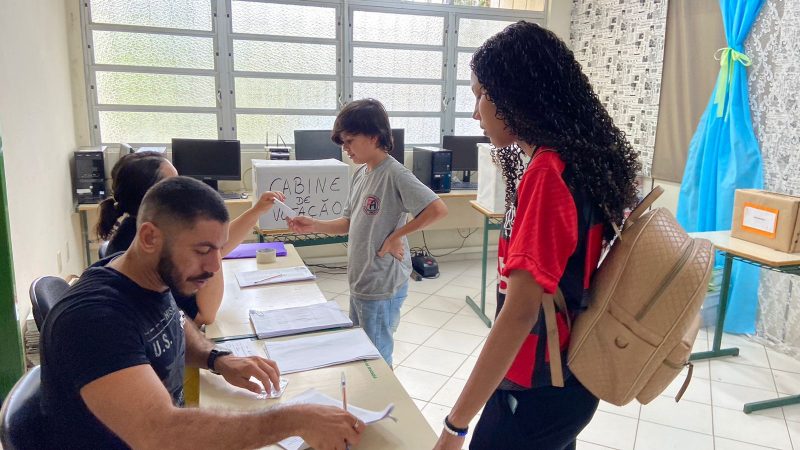 Foto mostra mesa eleitoral com dois mesários e dois estudantes pegando as cédulas de votação na escola EEB Padre Anchieta