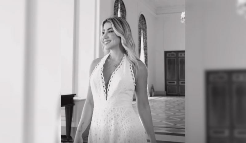 Bruna Olivo, empresária de Criciúma, em foto em pé, andando com um vestido branco e sorrindo. Ela é loira e usa um vestido com decote rendado profundo