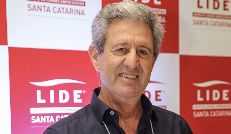 Homem branco de cabelos grisalhos sorrindo e posando em frente a um painel nas cores vermelho e branco. Ele é um dos bilionários catarinenses na lista da Forbes
