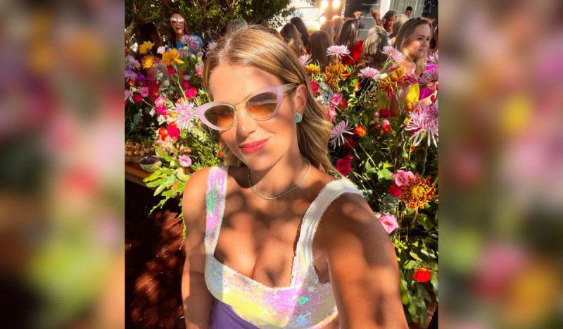 Lia Schlikmann, herdeira de SC. Na foto, mulher loira de óculos de sol fazendo selfie e ao fundo uma mureta de flores multicoloridas