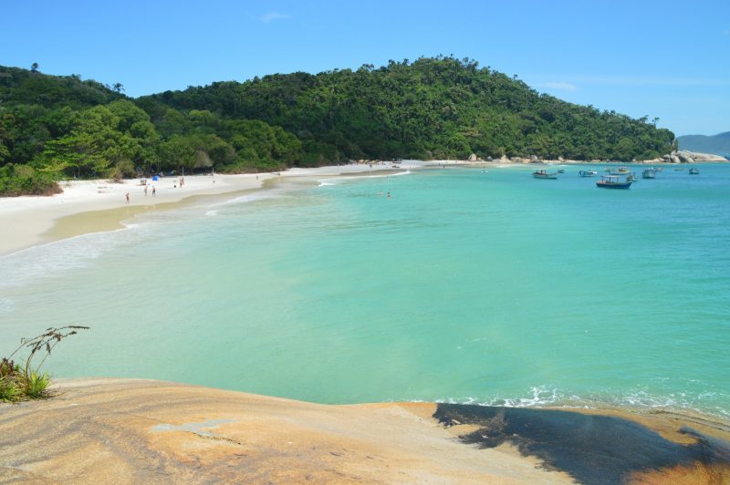 Foto da Ilha do Campeche. No centro da foto, o mar de cor azul-esverdeado. Ao fundo e no canto esquerdo a areia muito branca, cercada por um morro verde. No lado direito aparecem alguns pequenos barcos no mar. 