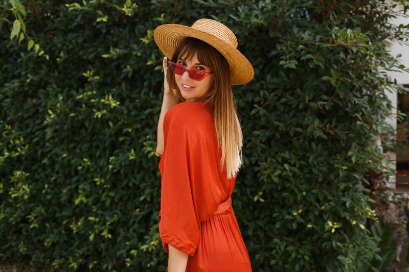 mulher com roupa laranja, cabelo liso na metade das costas, com as pontas loiras, com chapéu e óculos de sol vermelho na frente de uma árvore 