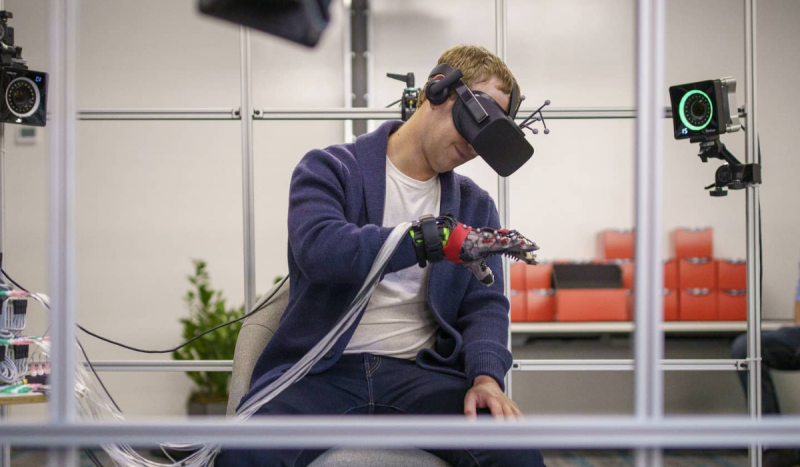 Mark Zuckerberg - Na foto, homem loiro usando óculos de realidade virtual e braço biônico, sentado em um ambiente que remete a um cenário virtual.