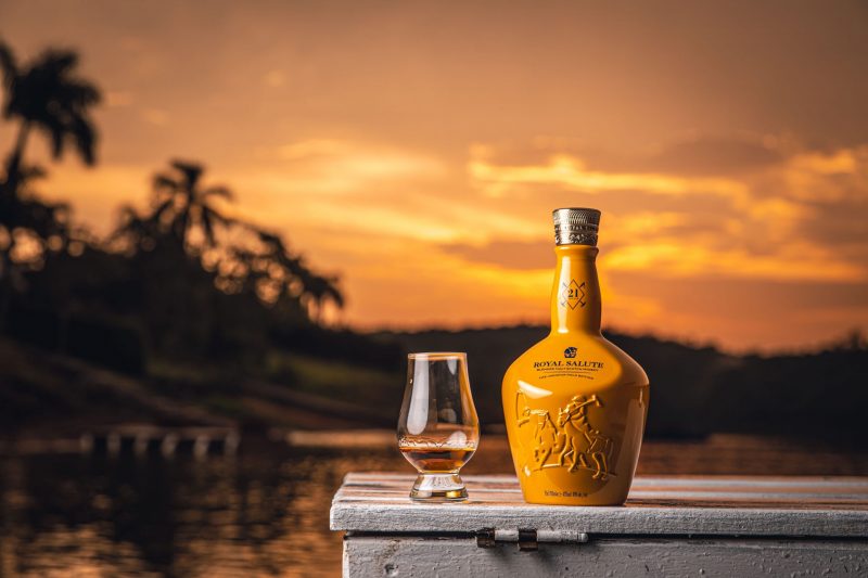Whisky Royal Salute ao lado de um copo em frente a um lago durante o entardecer