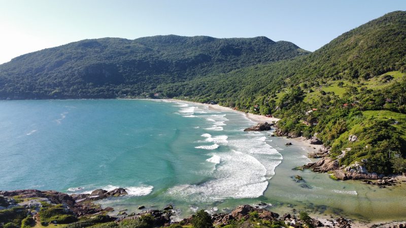 Foto da Praia de Matadeiro, também procurada para surfe em Florianópolis. Na parte inferior aparecem algumas rochas. No meio da foto o mar azul claro e no canto direito e parte superior, morros de vegetação verde, que cercam o mar. 