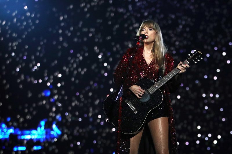 Taylor Swift, aparece de roupa vermelha brilhosa cantando em microfone com violão preto