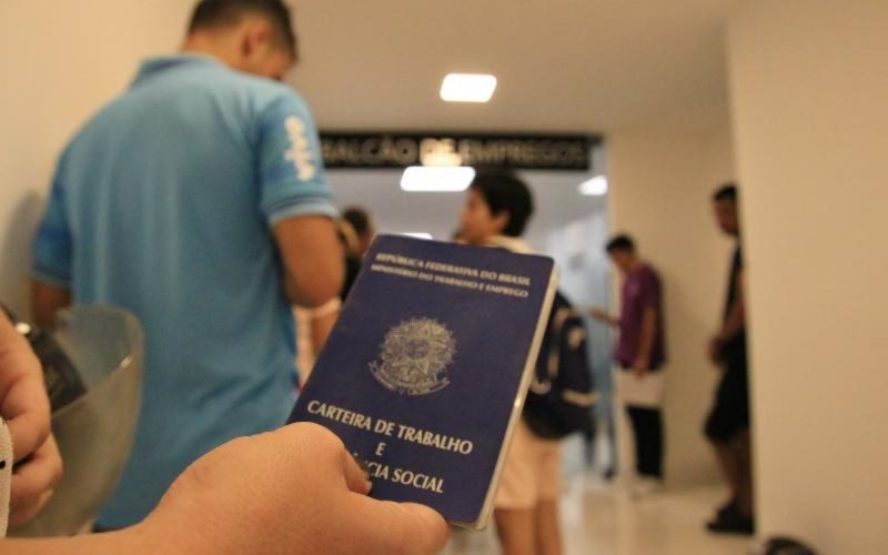 Foto mostra pessoas de pé em uma sala e alguém segurando uma carteira de trabalho