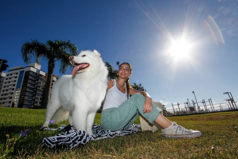 Giovanna De Carli, 17 anos, sentada sobre uma canga na grama, ao lado de um cachorro branco da raça samoieda, com o típico sol do verão ao fundo