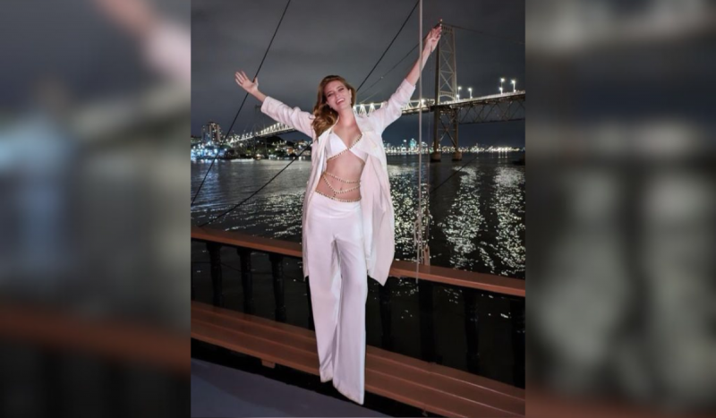 Mulher loira com traje branco posa sorrindo e de braços abertos em frente a uma ponte de metal