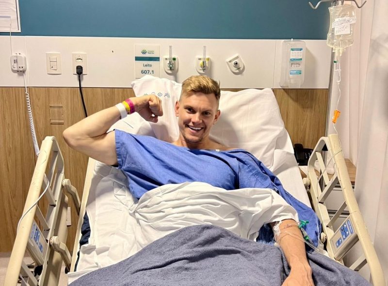 Douglas, deitado em uma cama de hospital, sorri após cirurgia