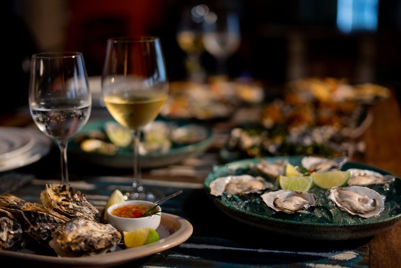 Foto de uma mesa cheia de pratos com ostras, em um restaurante de Florianópolis. Os dois primeiros pratos mostram ostras gratinadas e in natura. Ao deles, duas taças com vinho e água. Os demais pratos sobre a mesa aparecem desfocados ao fundo. 