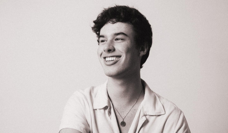 Fotógrafo de Florianópolis - Na foto, jovem branco de cabelos pretos e curtos sorri olhando para o lado esquerdo da foto. Ele usa camisa branca e um colar com pingente circular. A foto está com filtro preto e branco granulado.