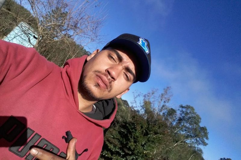 Dautiele Gonçalves da Silva, vítima do acidente na BR-280, usa moletom vermelho e um boné preto. Tem barba na cor castanha e é branco. Ao fundo, árvores e um céu azul