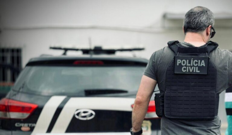 Policial civil de costas usando colete da PC e no fundo da foto aparece um carro preto