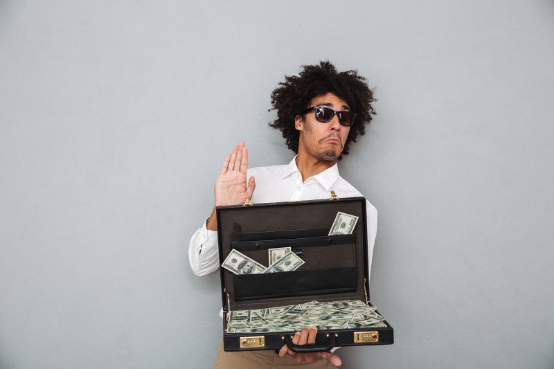 homem com cabelo black power, de óculos escuro, camisa social branca e calça marrom; ele está segurando uma maleta preta cheia de dólares 