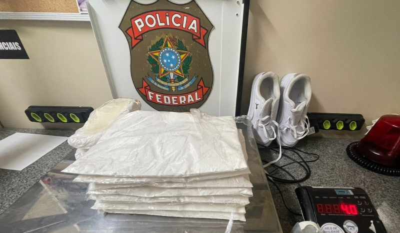 Foto mostra pacotes de cocaína em cima de uma balança de precisão. Na foto também aparece um par de tênis brancos e a insígnia da Polícia Federal