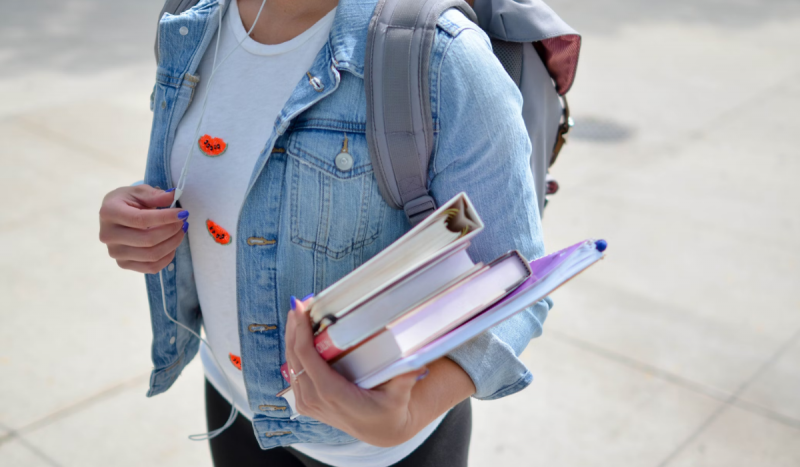 Menina usando blusa branca e jaqueta jeans segurando livros no braço esquerdo