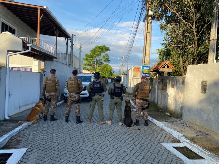 Três policias militares e dois policiais civis posam para foto de costas numa rua da Palhoça. Há dois cachorros acompanhando os policiais.
