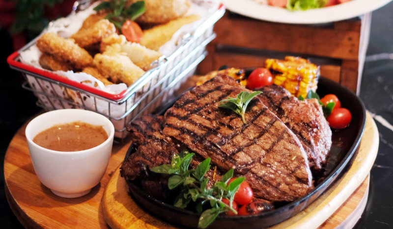 Peça de picanha assada em cima de travessa. Ao lado, pote pequeno de molho e empanados