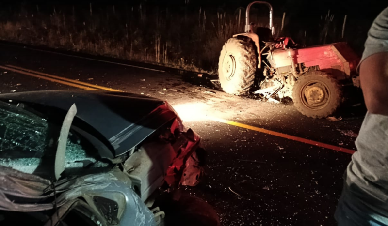 Carro cinza destruído em frente a trator na pista após acidente