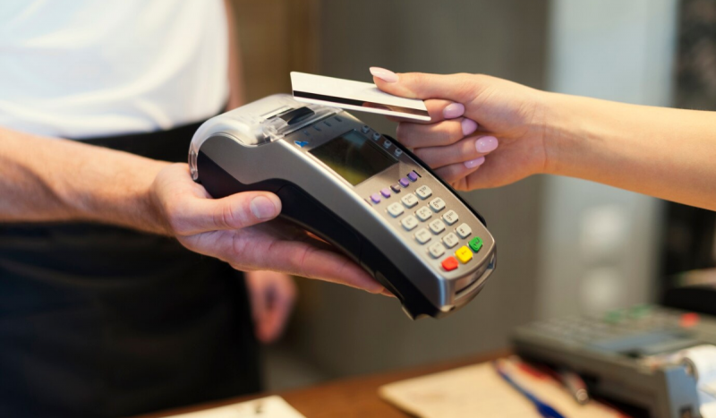 Mão feminina com unhas na cor rosa segurando cartão de crédito passando na maquininha de cartão