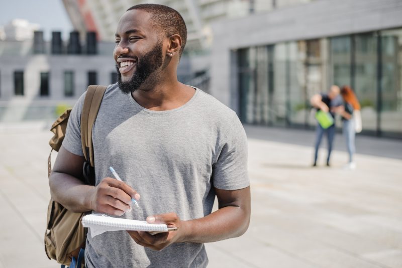 Estudante carregando mochila nas costas, com caderno e caneta na mão