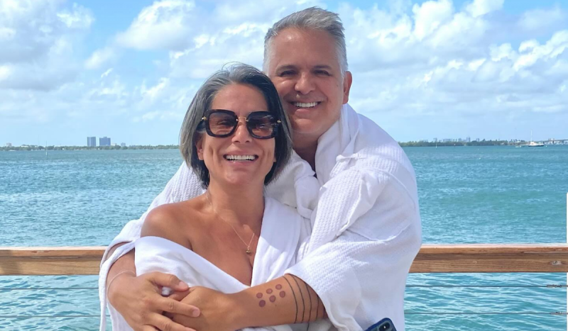 Glória Pires e o marido abraçados - Na foto, homem branco e mulher branca de óculos escuros posam de roupão branco abraçados em frente ao mar