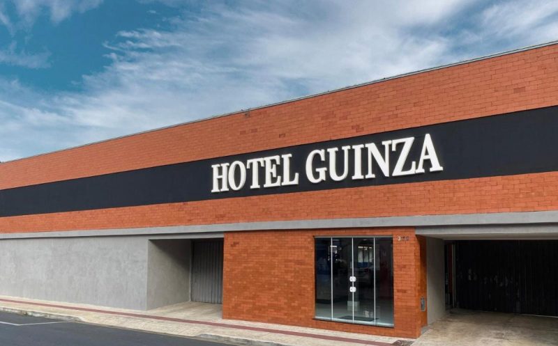 Fachada do Guinza, um hotel barato em Balneário Camboriú. A fachada é de tijolos marrom com uma listra preta de fora a fora por cima da qual está escrito o nome do hotel em letras brancas grandes.