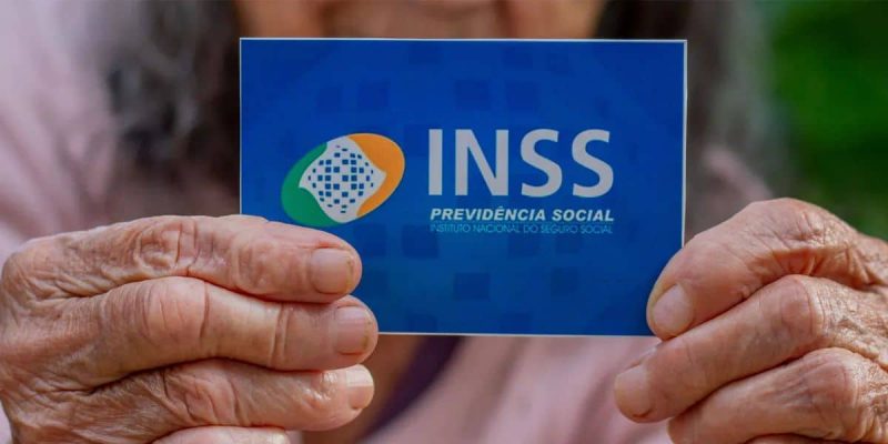 Foto mostra duas mãos de pessoa idosa segurando cartão azul escrito 'INSS - Previdência Social'