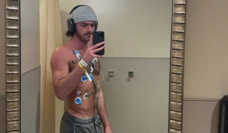 Jovem branco usando touca cinza na cabeça, posando em frente ao espelho tirando foto com o celular. Ele usa diversos eletrodos no corpo em razão do uso exagerado de vape