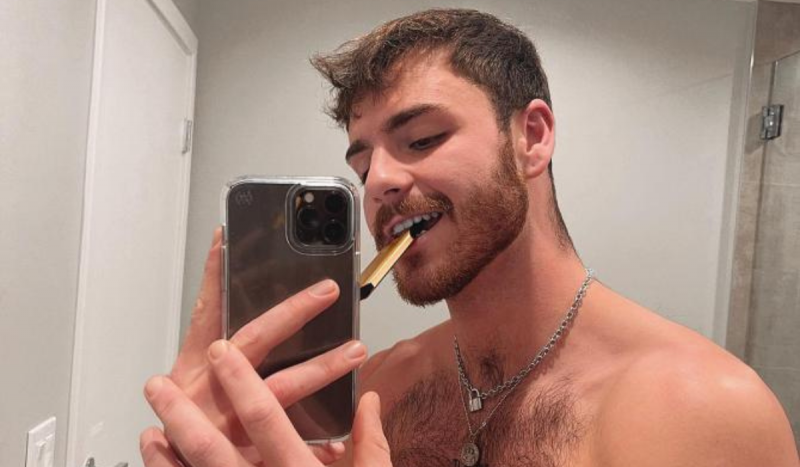 Homem branco posando sem camisa em frente ao espelho com um cigarro eletrônico dourado no meio dos dentes