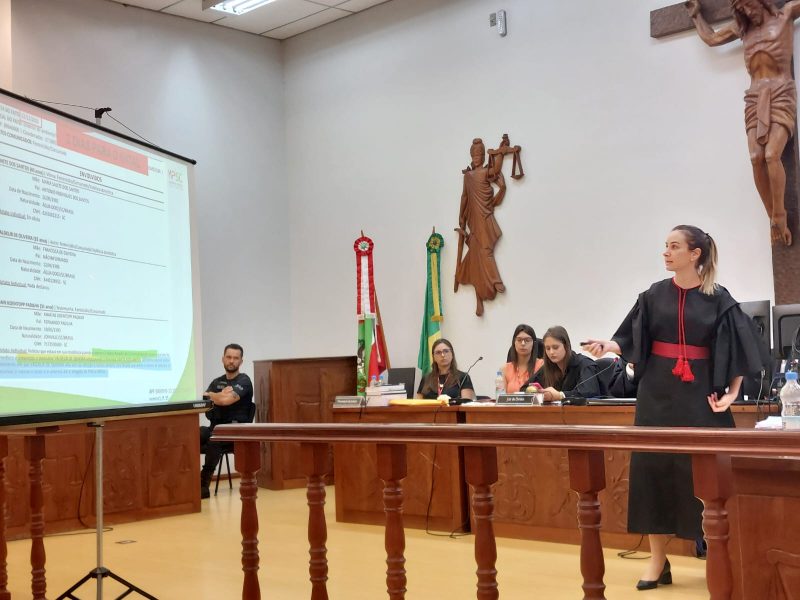 Júri do homem que matou a mulher grávida em Ibicaré, na imagem aparece a promotora de Justiça mostrando em um telão as informações sobre o processo. Ela está na sala do fórum da comarca de Ibicaré e aparece em pé de frente para os jurados. atrás dela aparecem mulheres sentadas e observando a explanação da promotora. 