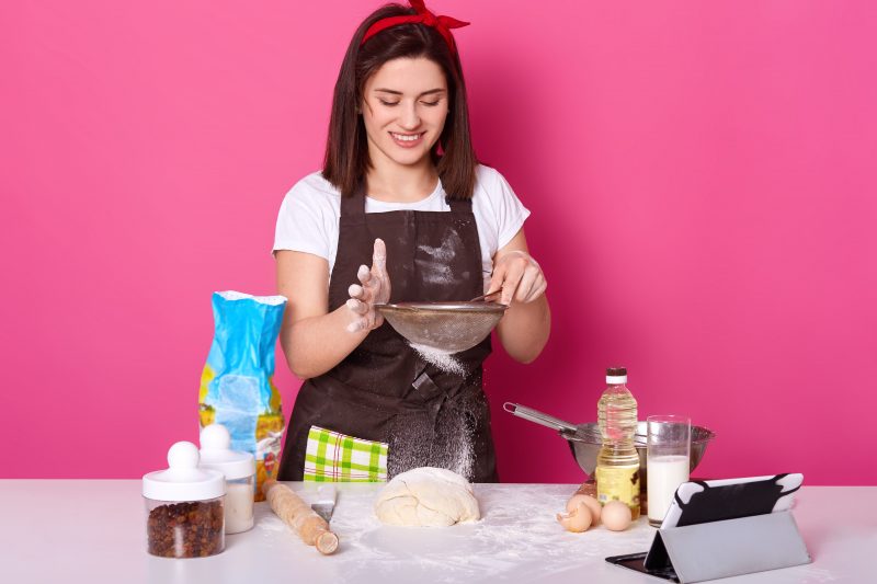 Retrato de uma cozinheira colocando farinha na peneira na torta meio pronta. Morena jovem posa isolada sobre fundo rosa brilhante. Conceito de cozimento e culinária.