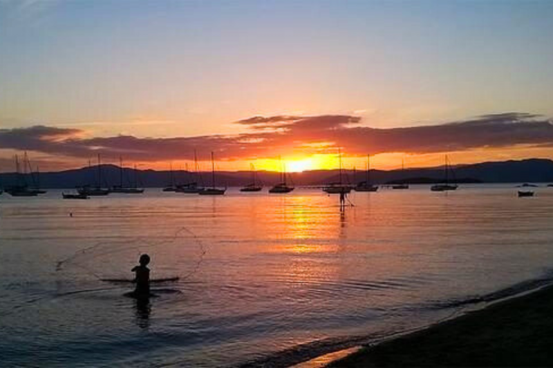 Foto do mar com uma criança jogando uma rede de pesca. Ao fundo, vários barcos parados. Eles e a criança estão em contraluz, escuros. No céu o pôr do sol. Trata-se da Praia de Santo Antônio de Lisboa, em Florianópolis. 