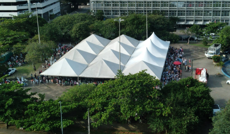 Foto aérea mostra tenda branca com pessoas reunidas embaixo. Ao redor muitas árvores aparecem vistas de cima. Foto mostra servidores decidindo por greve na educação de SC