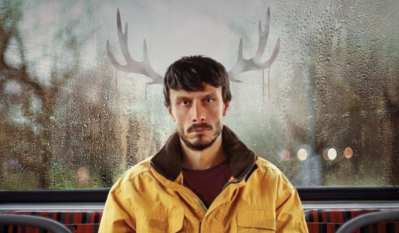 Cartaz da série Bebê Rena mostra homem sentado em frente a janela de vidro usando casaco amarelo e um chifre de rena