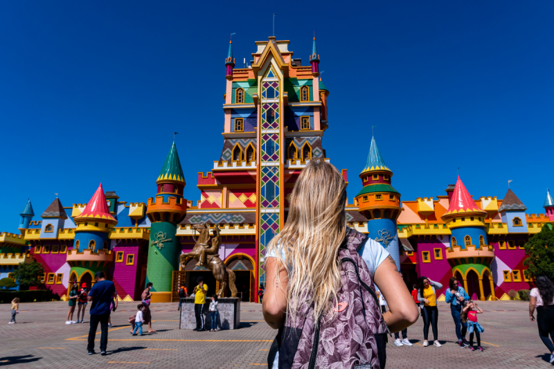 Foto de frente do Castelo das Nações, um prédio todo colorido e com várias torres onde fica a bilheteria e a entrada do Beto Carrero World. À frente dele há várias pessoas, de diferentes características físicas, espalhadas. De costas para foto e em destaque há uma jovem branca, de cabelos compridos e loiros com uma mochila em tom de roxo. 