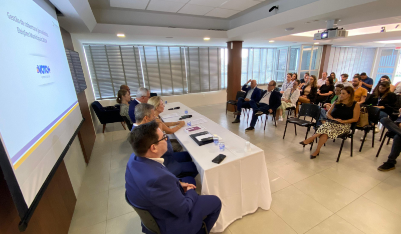 Pessoas sentadas em plateia em frente à mesa longa com toalha branca, onde estão sentadas pessoas do TRE-SC, para falar das eleições 2024