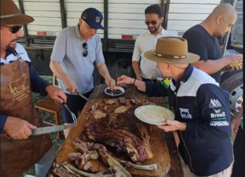 Na imagem, aparece várias pessoas degustando uma costela assada durante almoço do CTG Os Praianos