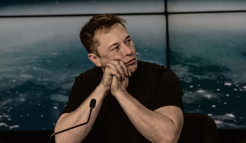 Elon Musk em entrevista coletiva com as mãos cruzadas apoiadas no queixo