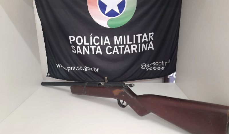 Espingarda em cima de mesa branca e ao fundo uma bandeira preta com a insígnia da polícia militar de Santa Catarina