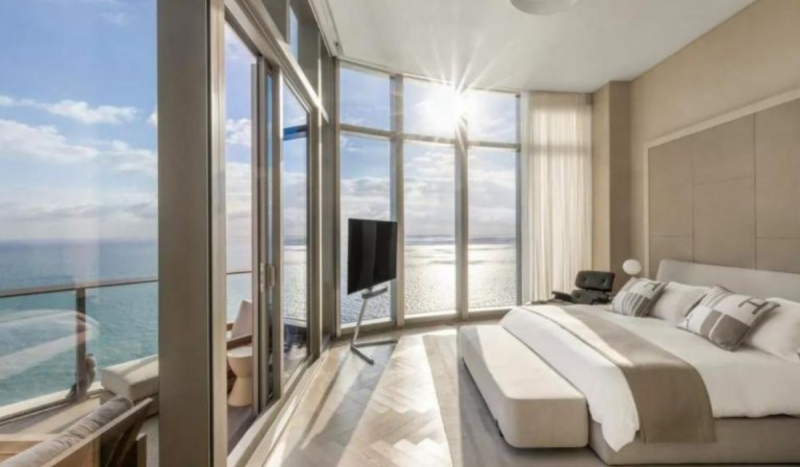 Foto de quarto em cobertura com vista panorâmica para o mar. Foto mostra janelas do chão ao teto, com sol incidindo. 