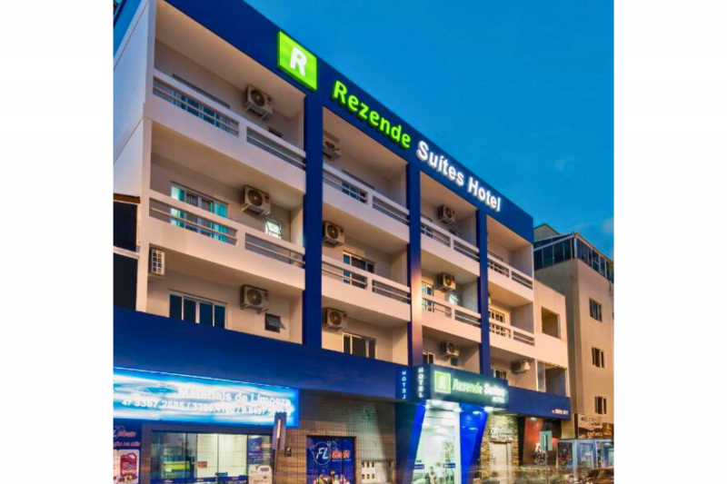 Fachada do Rezende Suítes, um hotel barato em Balneário Camboriú. O hotel tem três andares com grandes sacadas. Ele mistura as cores branca e azul. O nome do hotel está escrito em verde e branco. 